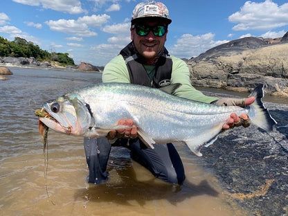 Fisherman in a cap holding a big payara caught on a hand-tied Payara Dragon Fly with boulders in the background